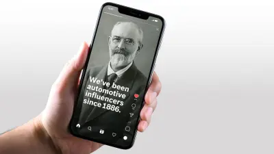 Detail ruky držící chytrý telefon zobrazující příspěvek ze sociálních médií s historickým portrétem Roberta Bosche a textem „Jsme automobilovými influencery od roku 1886.“
